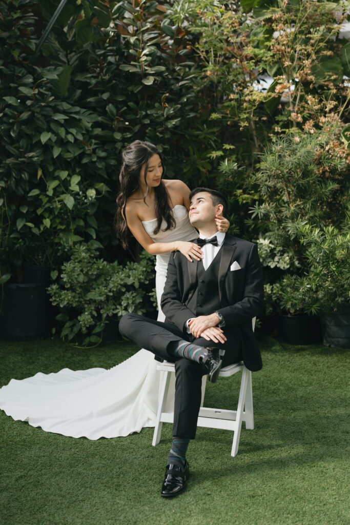 Groom sitting in a chair while the bride has her arms around him.