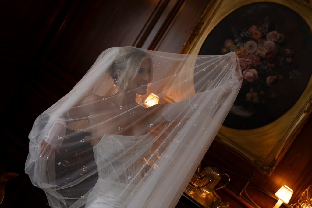 Bride smiling as she holds up her hand under her veil in an antique room at the Aldredge House.