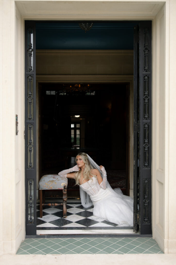 Bride sitting on the floor while leaning on a piece of furniture, adjusting her veil.